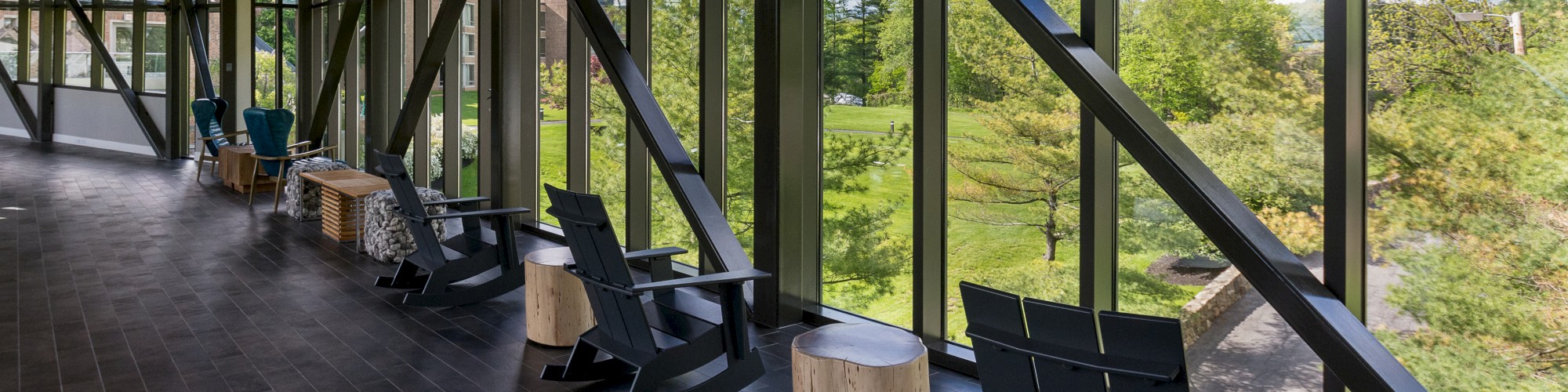 A sunlit lounge area with black rocking chairs and small wooden tables facing large windows providing a view of greenery outside.