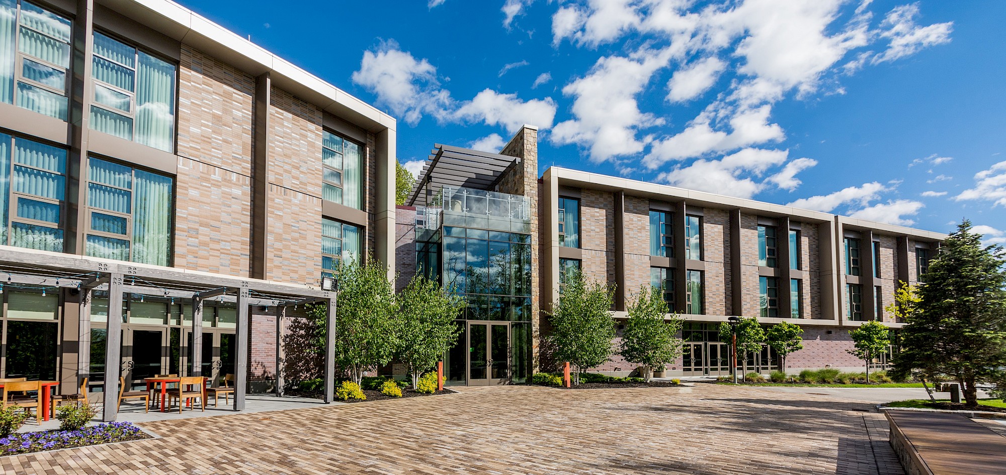 A modern two-story building with large windows, surrounded by trees and a paved pathway under a bright blue sky with scattered clouds.