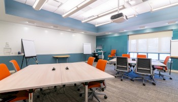 This image depicts a modern conference room with tables, orange chairs, a projector, whiteboards, and natural lighting from large windows.