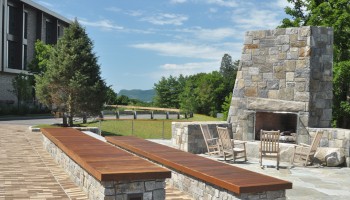 The image shows an outdoor setting with a stone fireplace, wooden benches, and rocking chairs on a patio. Trees and a building are in the background.