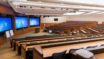 An empty lecture hall features multiple rows of tiered seating, three large screens at the front, several whiteboards, and a podium with a computer.