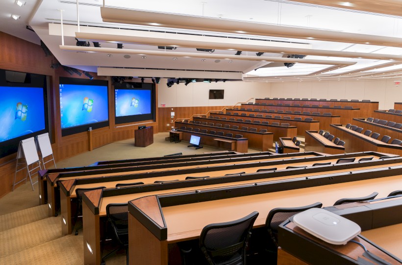 An empty lecture hall features multiple rows of tiered seating, three large screens at the front, several whiteboards, and a podium with a computer.