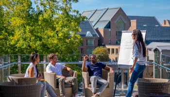 A group is having an outdoor meeting on a terrace with a flip chart. They're sitting in wicker chairs, surrounded by trees and buildings.