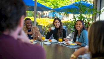 A group of people sitting at a table outside, enjoying drinks and food. They appear to be having a conversation and smiling.