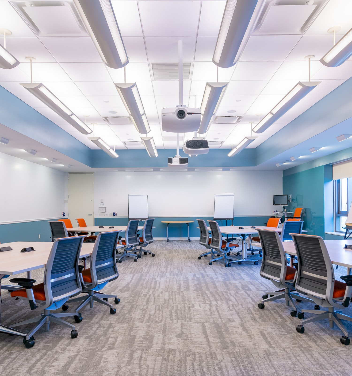 A modern conference room with U-shaped tables, ergonomic chairs, whiteboards, and ceiling-mounted projectors, featuring bright lighting.