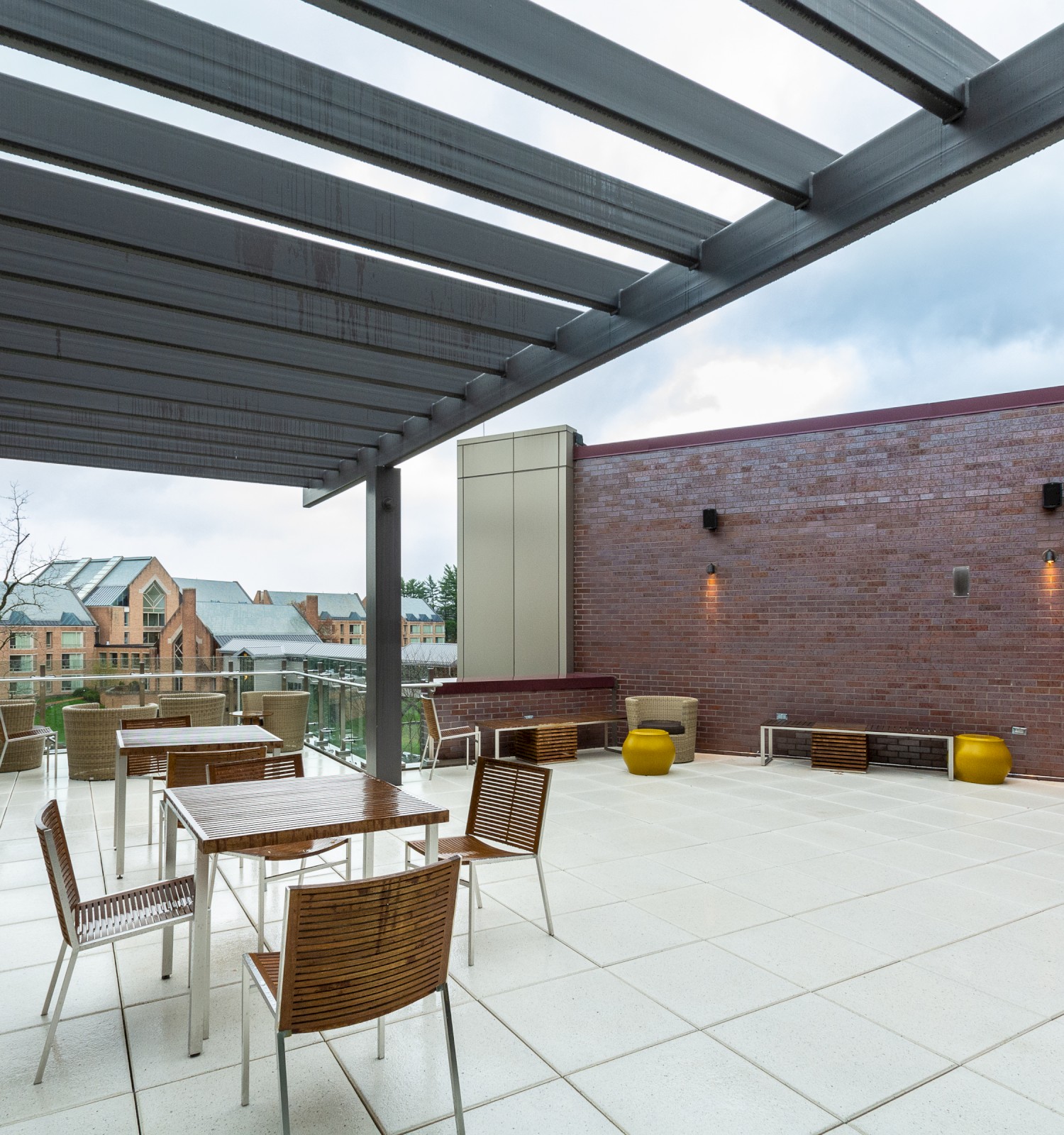 An outdoor patio area with pergola, wooden chairs, and metal tables, surrounded by brick walls and overlooking a scenic background with buildings in the distance.