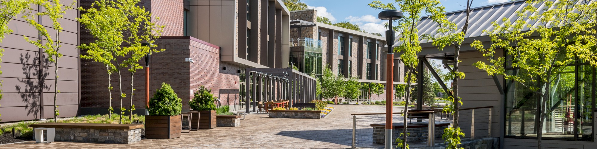 A modern courtyard with brick pathways, various plants, benches, trees, contemporary buildings, and a clear blue sky with scattered clouds.