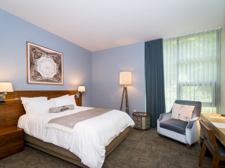 A modern bedroom with a double bed, wall art, large window with curtains, armchair, bedside tables, and floor lamp, featuring neutral colors.