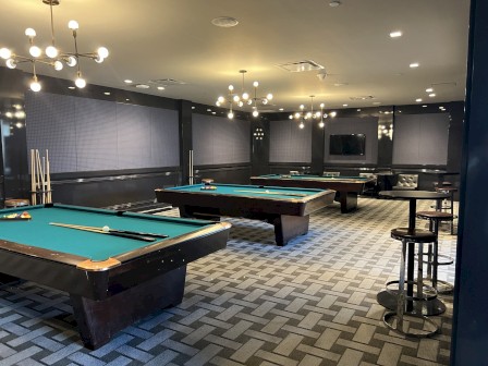 The image shows a stylish billiards room with several pool tables, modern light fixtures, stools, and a flat-screen TV mounted on the wall.