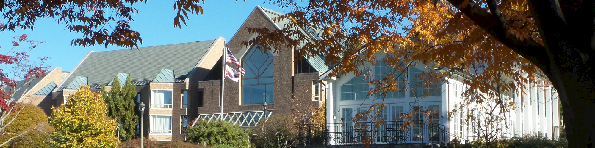 A building complex with peaked roofs, surrounded by trees with autumn foliage, and an open grassy area in the foreground, under a clear sky.