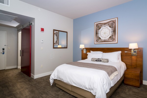 A cozy hotel room with a neatly made bed, modern lighting, a red sliding door, a mirror, and a framed artwork on the blue accent wall.