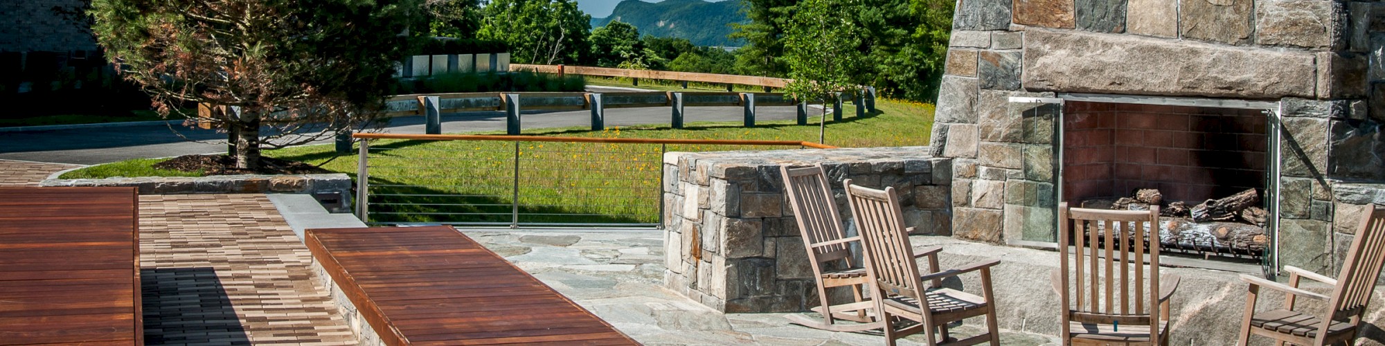 An outdoor patio has wooden benches, stone fireplace, and rocking chairs, surrounded by green trees and a blue sky in the background.