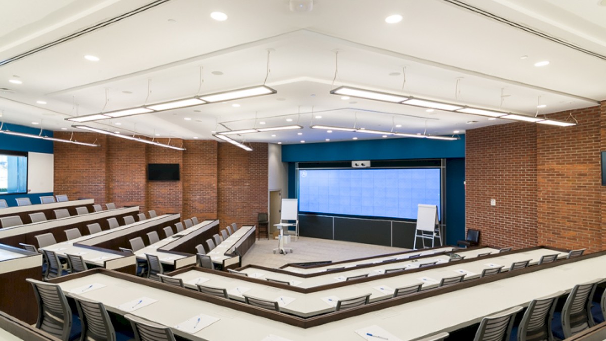 This image shows an empty lecture hall with rows of seats, a large screen at the front, a whiteboard, and a podium. The walls are brick.