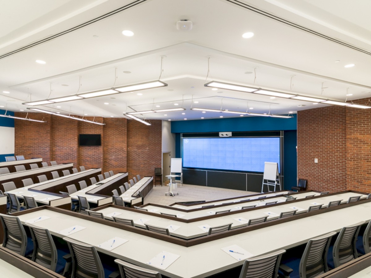 This image shows an empty lecture hall with rows of seats, a large screen at the front, a whiteboard, and a podium. The walls are brick.