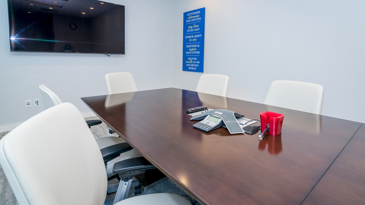 A conference room with a wooden table, white chairs, a wall-mounted television, and some items on the table, including a red cup.