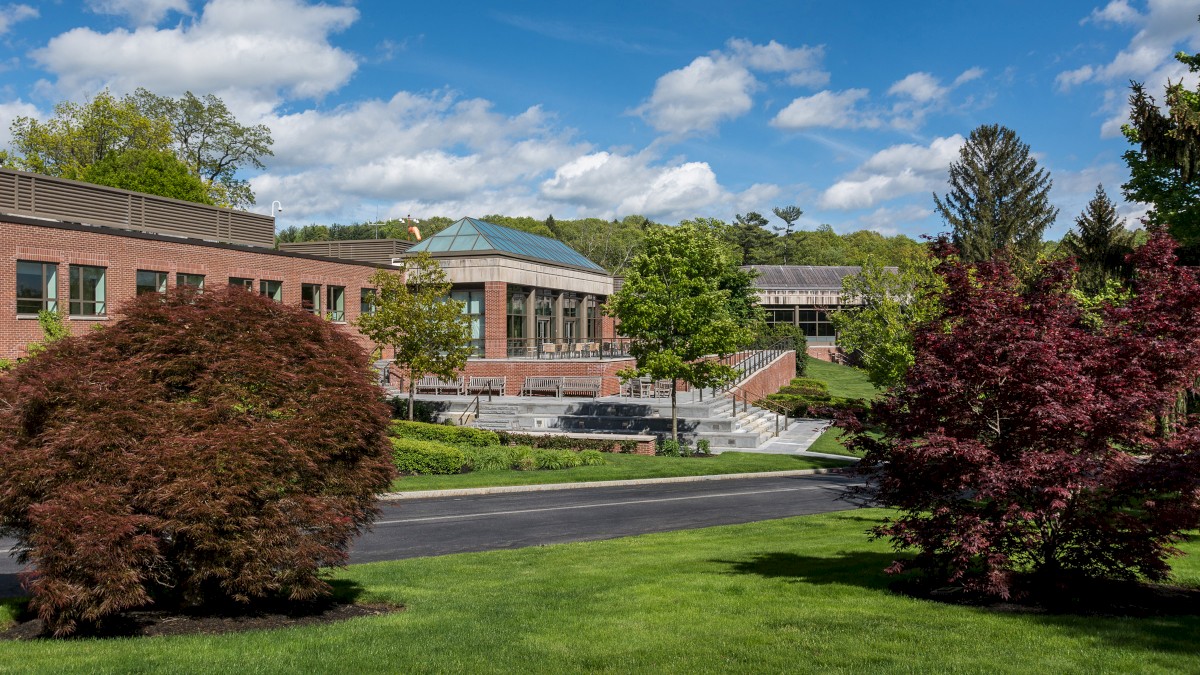 The image shows a picturesque, landscaped campus with green lawns, trees, and a modern building against a backdrop of a partly cloudy sky.