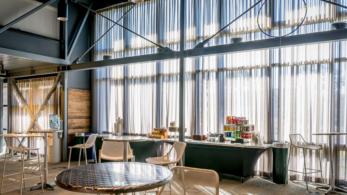 This image shows a stylish, sunlit cafeteria or break room with modern tables, chairs, and a refreshment area offering drinks and snacks.