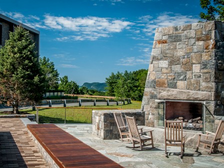 An outdoor seating area with wooden benches, stone fireplace with rocking chairs, and a scenic view of greenery on a clear day, always ending the sentence.