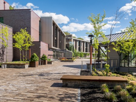 A modern courtyard with buildings, trees, benches, and a brick walkway under a blue sky with clouds.