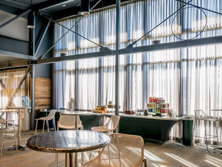 The image shows a modern interior space with tables, chairs, large windows with curtains, and a refreshment area with various beverages and snacks.