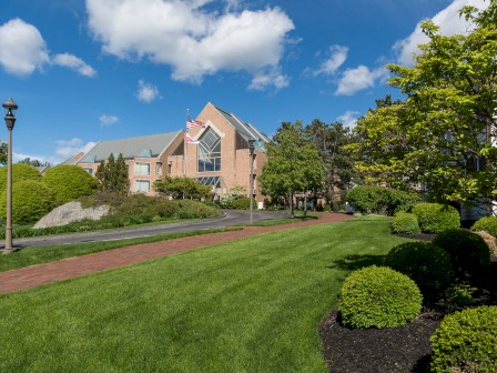 A well-maintained campus with a brick building, lush green lawn, and trimmed bushes, under a clear blue sky with fluffy clouds.