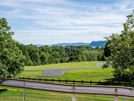 The image shows a scenic landscape with a paved path winding through a lush green park, surrounded by trees and distant hills under a partly cloudy sky.