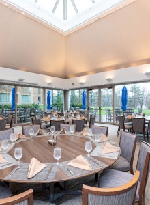 The image shows an elegant restaurant with round tables set with glasses and napkins, large windows, and a skylight providing natural light.