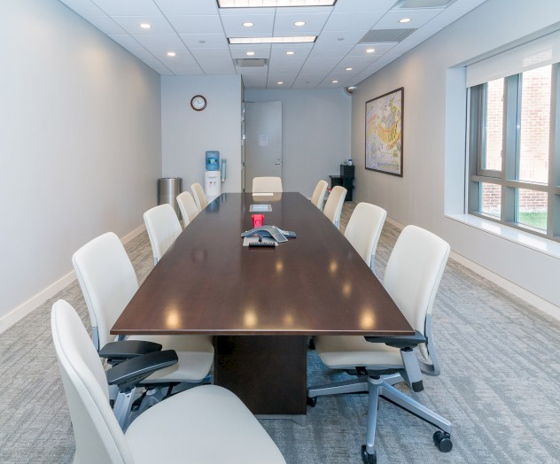 The image shows a modern conference room with a long wooden table, white office chairs, a wall-mounted screen, and large windows on one side.