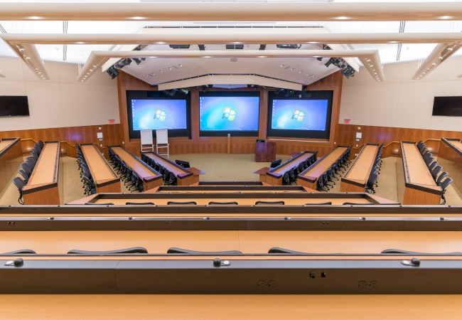 A modern lecture hall with tiered seating, multiple large screens displaying the Windows desktop screen, and a podium at the front of the room.