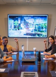 A group of four people are in a meeting room with notepads, a screen displaying an image, and a flipchart stand to the side.