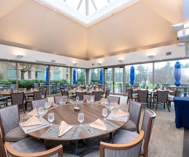 The image shows an elegant restaurant with round tables set with glasses and napkins, large windows, and a skylight providing natural light.