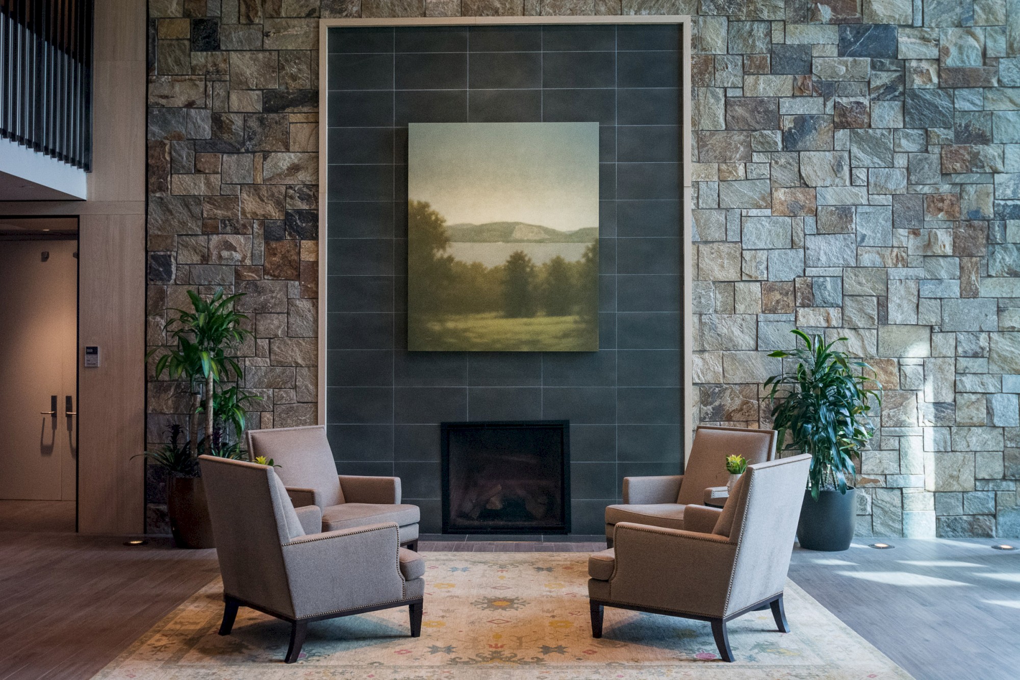 The image shows a cozy seating area with four armchairs around a fireplace, a painting above it, and surrounded by stone walls and green plants.