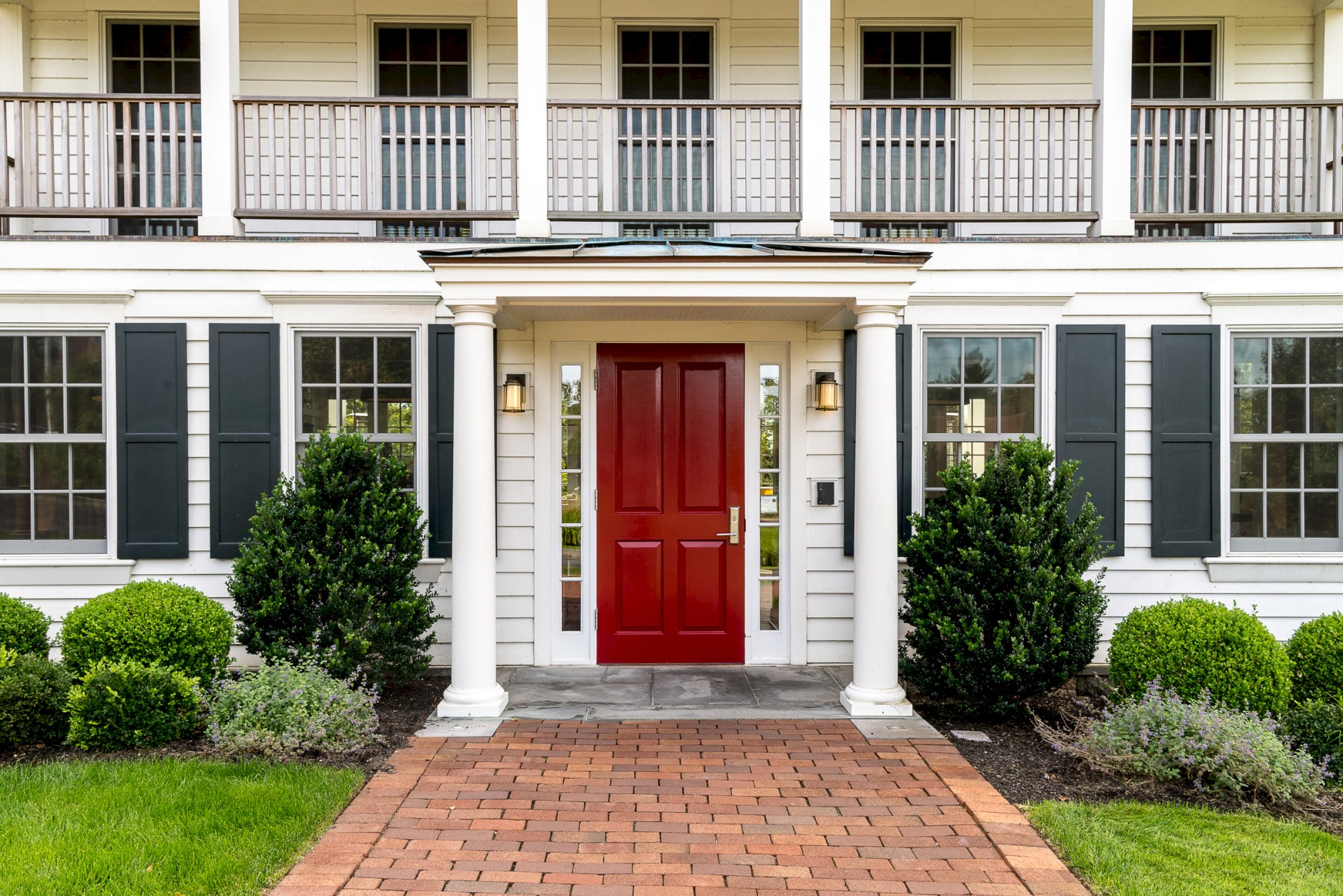 A two-story house with a red front door, white siding, black shutters, and a brick walkway, surrounded by green shrubs and well-kept landscaping.