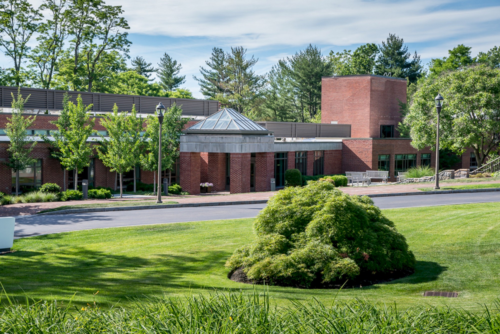 The image shows a brick building surrounded by lush green lawns and trees, under a partly cloudy sky with a well-maintained landscape.