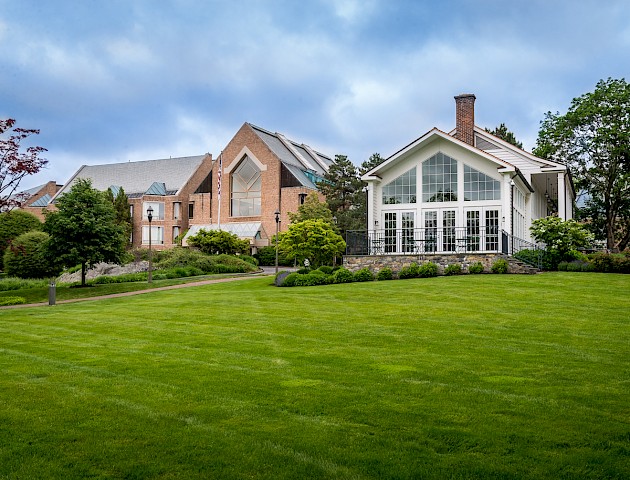 A large, well-manicured lawn with two adjacent buildings: a modern brick structure and a white house with large windows, against a cloudy sky.