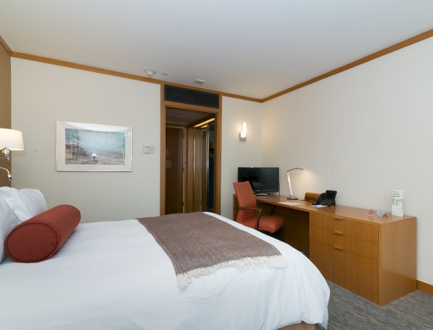 A neatly arranged hotel room with a bed, desk, chair, lamp, and artwork on the wall, along with a door leading to another space.