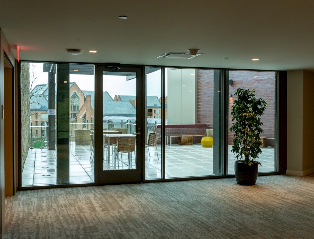 The image shows a modern indoor space with large glass windows opening to an outdoor patio, with potted plants and outdoor seating visible outside.