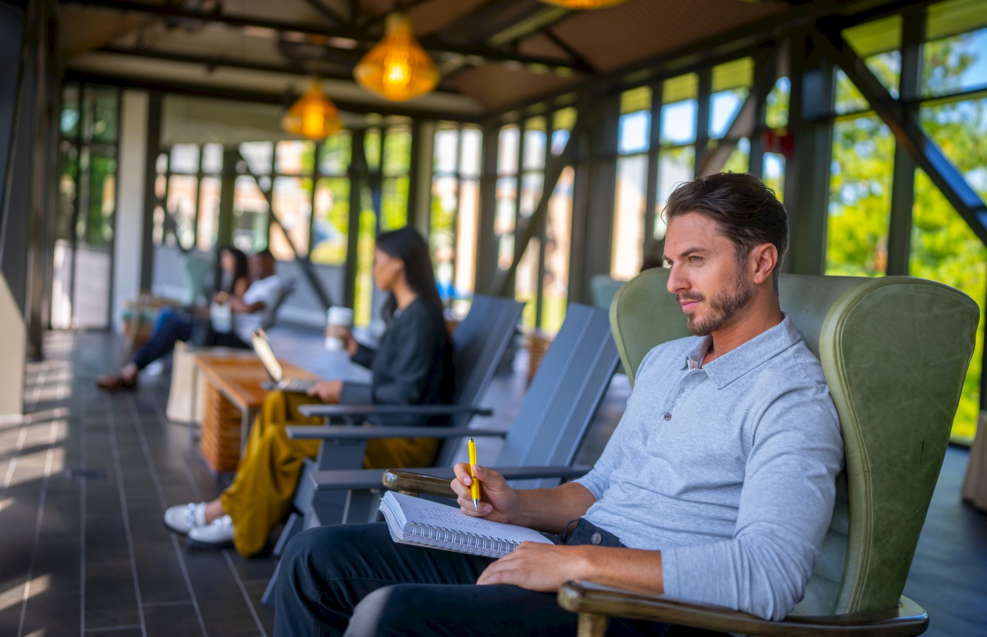 People are sitting in a bright indoor space, focusing on reading or writing. The room has large windows and warm lighting.