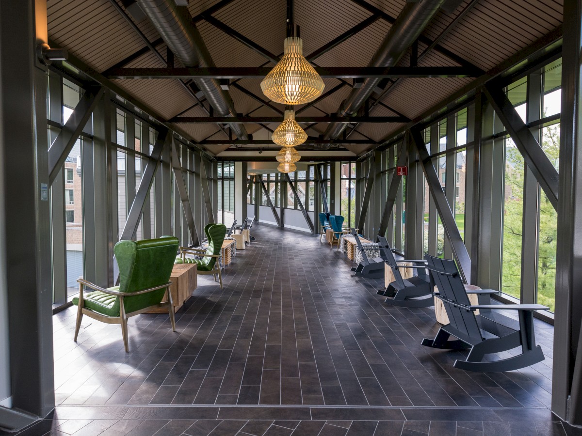 A long corridor with large windows, green and blue chairs, and hanging chandeliers. The floor is tiled, and light streams in from the windows.