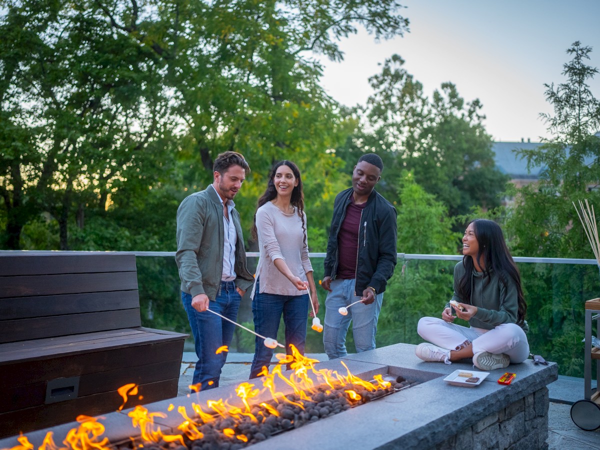 A group of friends roasting marshmallows over an outdoor fire pit, surrounded by trees and a relaxed atmosphere, enjoying the evening.