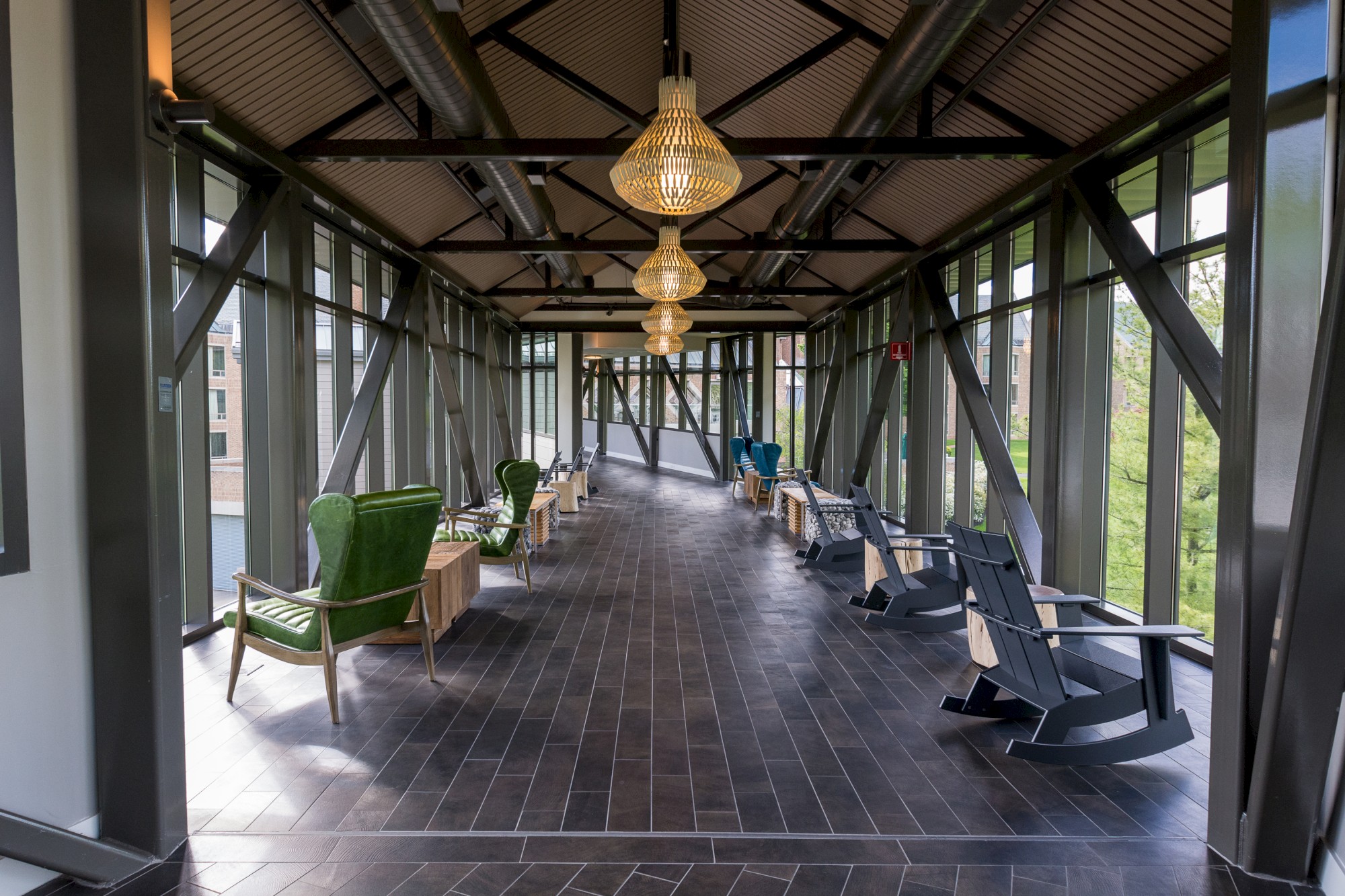 A modern, glass-walled hallway features stylish armchairs, rocking chairs, and hanging pendant lights, offering a scenic view with indoor comfort.