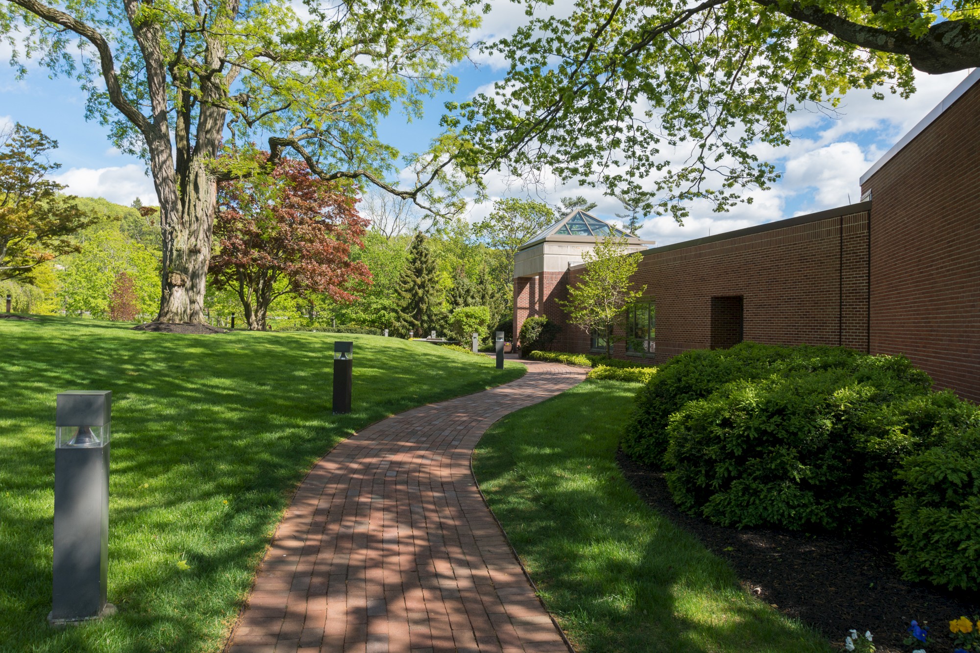 A brick pathway winds through a lush green garden, leading to a modern building with a glass roof, surrounded by trees and manicured hedges.