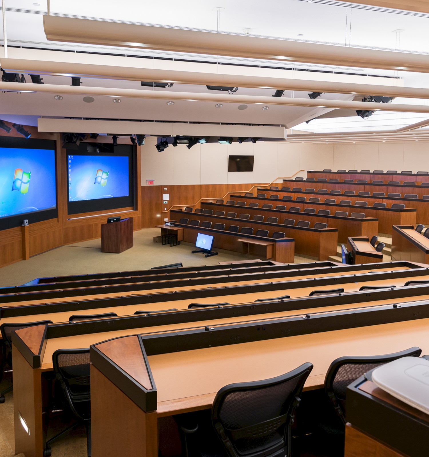 The image shows an empty lecture hall with multiple rows of seating, three large projection screens, and a podium at the front.