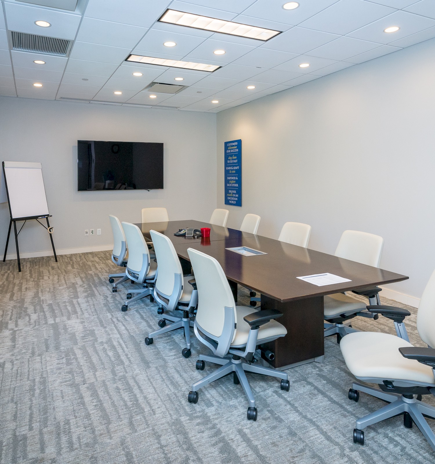 A modern conference room with a long table, eight chairs, a TV screen, a flip chart, and ample lighting.