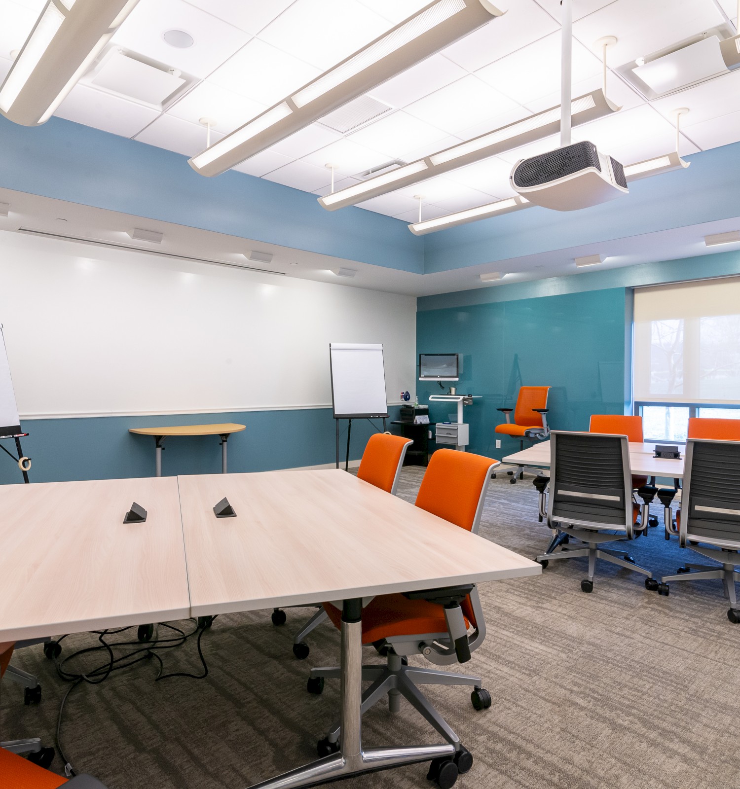 A modern conference room with tables, orange and black chairs, whiteboards, flip charts, a projector, and a large window is shown.