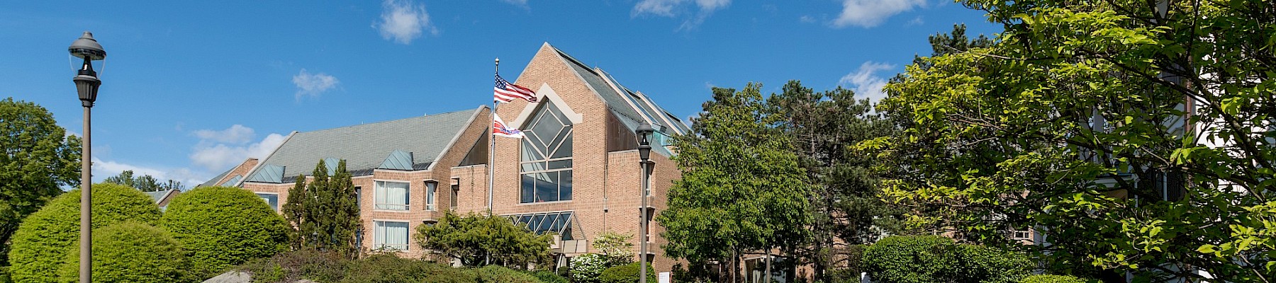 A red brick building with a flagpole, manicured lawns, and a path, under a blue sky with scattered clouds.