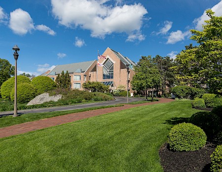 A red brick building with a flagpole, manicured lawns, and a path, under a blue sky with scattered clouds.