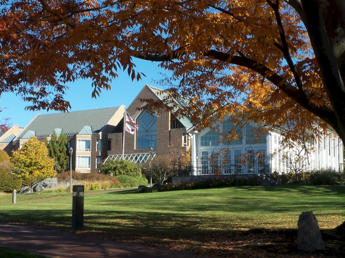 The image shows a campus building with fall foliage, a grassy lawn in the foreground, and a pathway leading towards the structure.