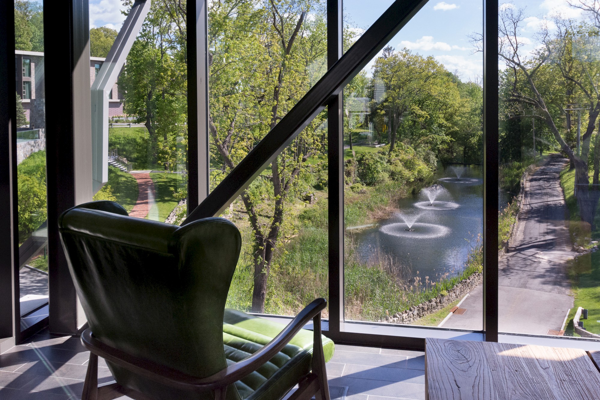 A green armchair by floor-to-ceiling windows overlooks a scenic view with a river, trees, and fountains in a serene outdoor setting.