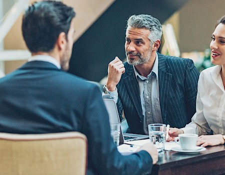 A group of four people, two men and two women, are in a discussion around a table with drinks and papers in a professional setting.